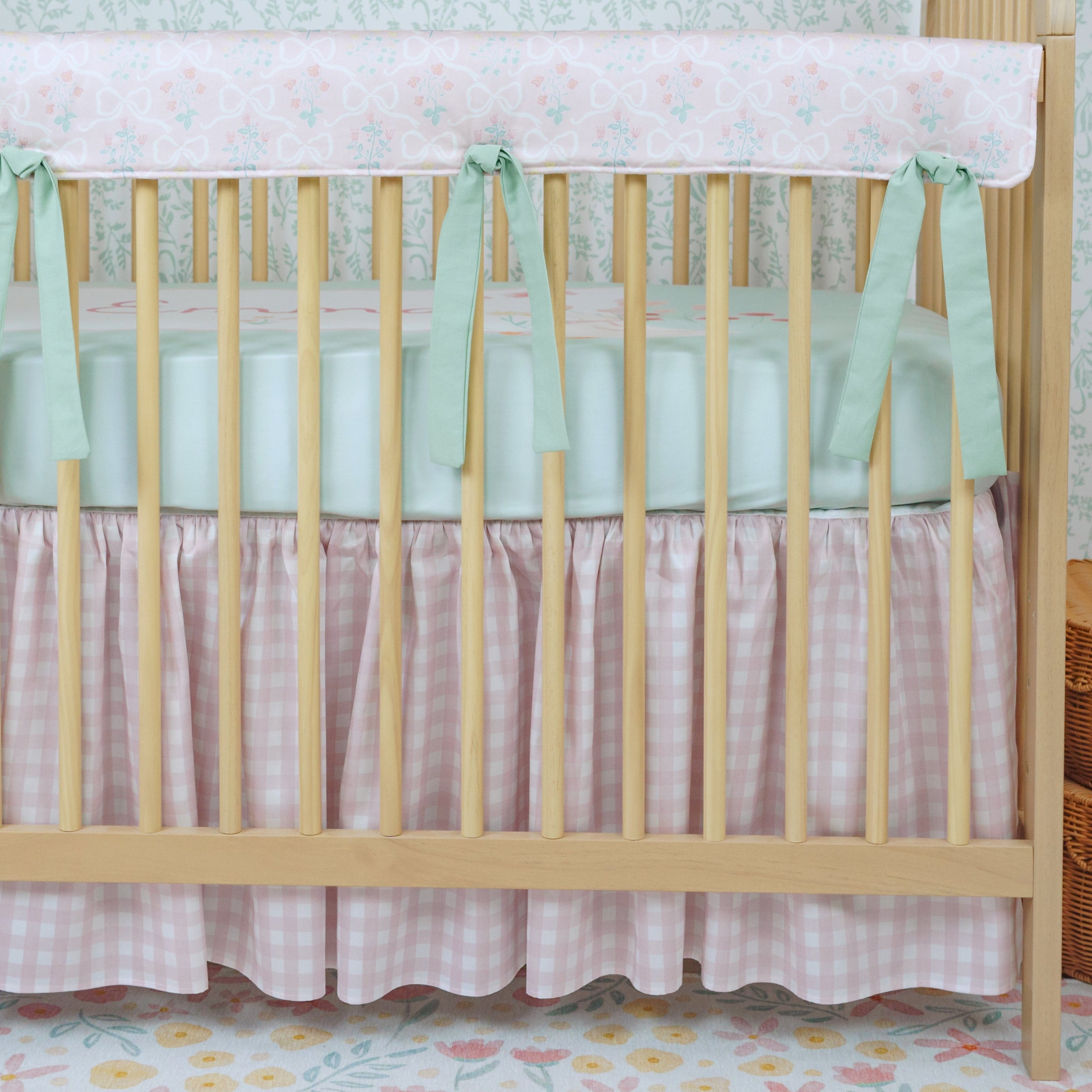 Wooden crib with white mattress and green bumper, light blue wall with floral wallpaper, wicker basket.