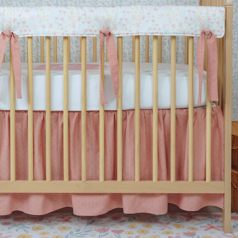 Wooden crib with pink bedding and white mattress in a room with floral wallpaper.
