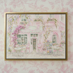 A framed art print depicting a whimsical pink cafe exterior with flowers and outdoor seating, mounted on a light pink floral wallpaper background.