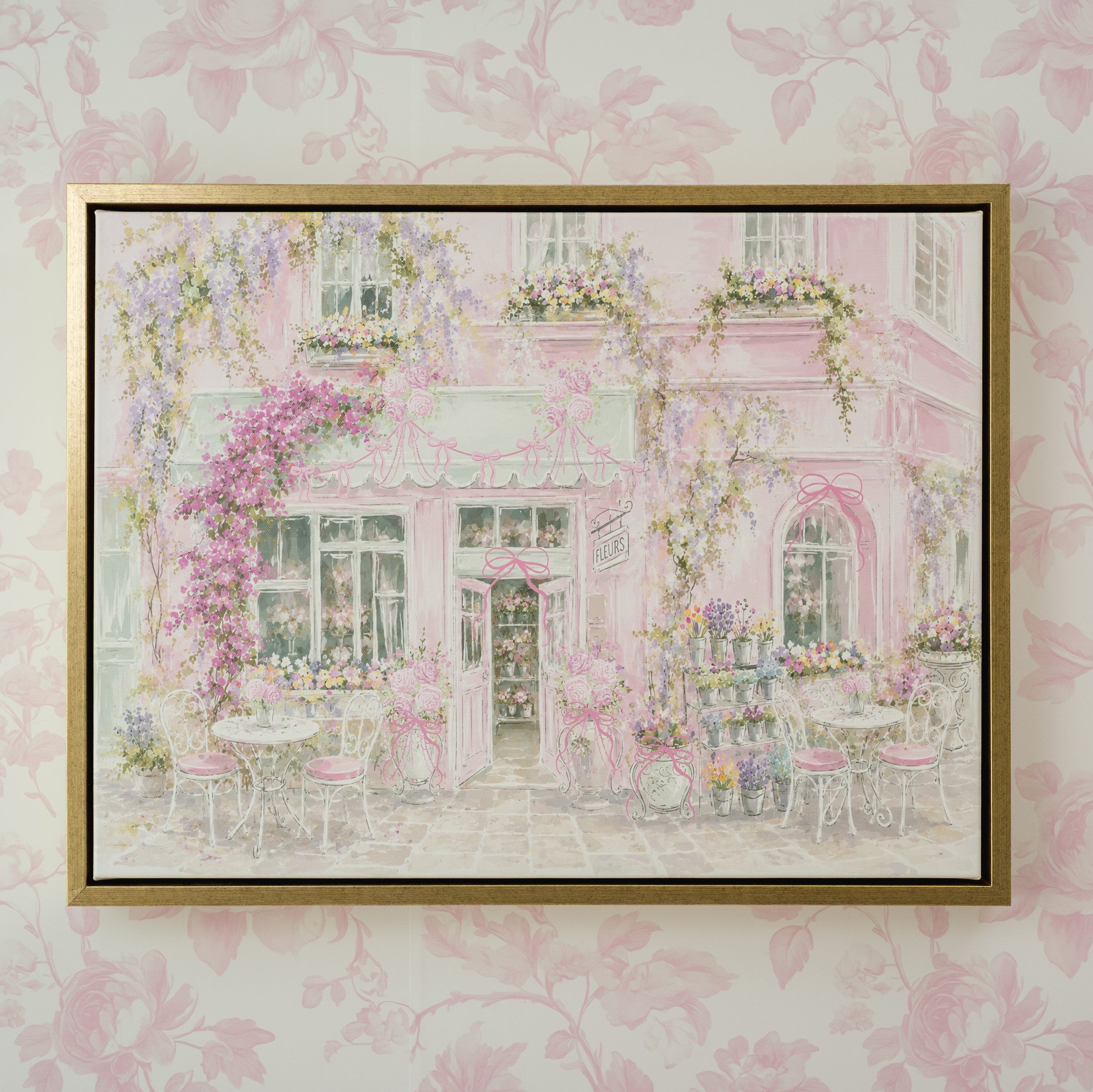 A framed art print depicting a whimsical pink cafe exterior with flowers and outdoor seating, mounted on a light pink floral wallpaper background.