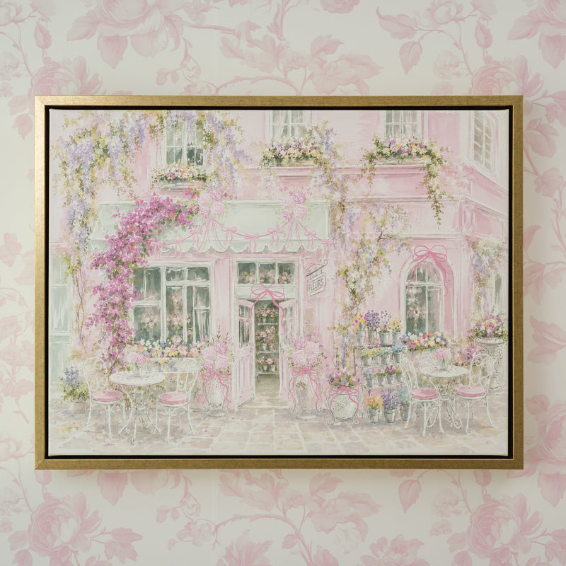 A framed art print depicting a whimsical pink cafe exterior with flowers and outdoor seating, mounted on a light pink floral wallpaper background.