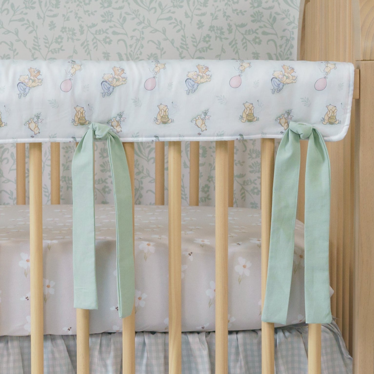 Wooden crib with floral crib sheet and green ribbons against a light blue plaid floor and patterned wall.