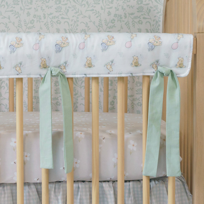 Wooden crib with floral crib sheet and green ribbons against a light blue plaid floor and patterned wall.