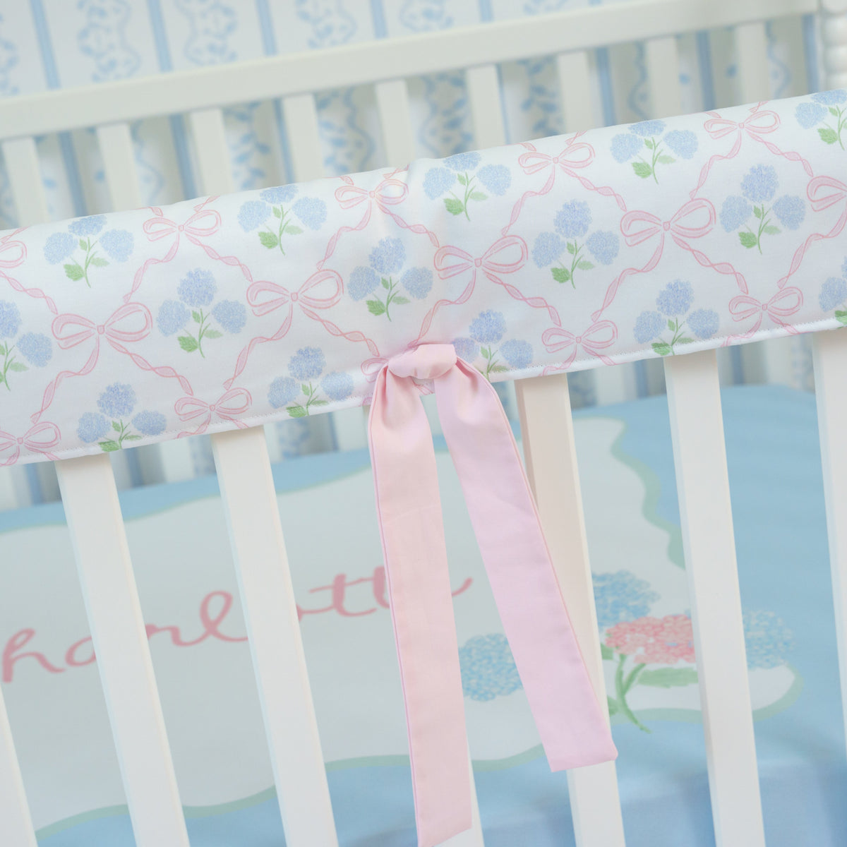 Crib with floral-patterned bumper and pink ribbon, on a light blue background