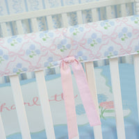 Crib with floral-patterned bumper and pink ribbon, on a light blue background