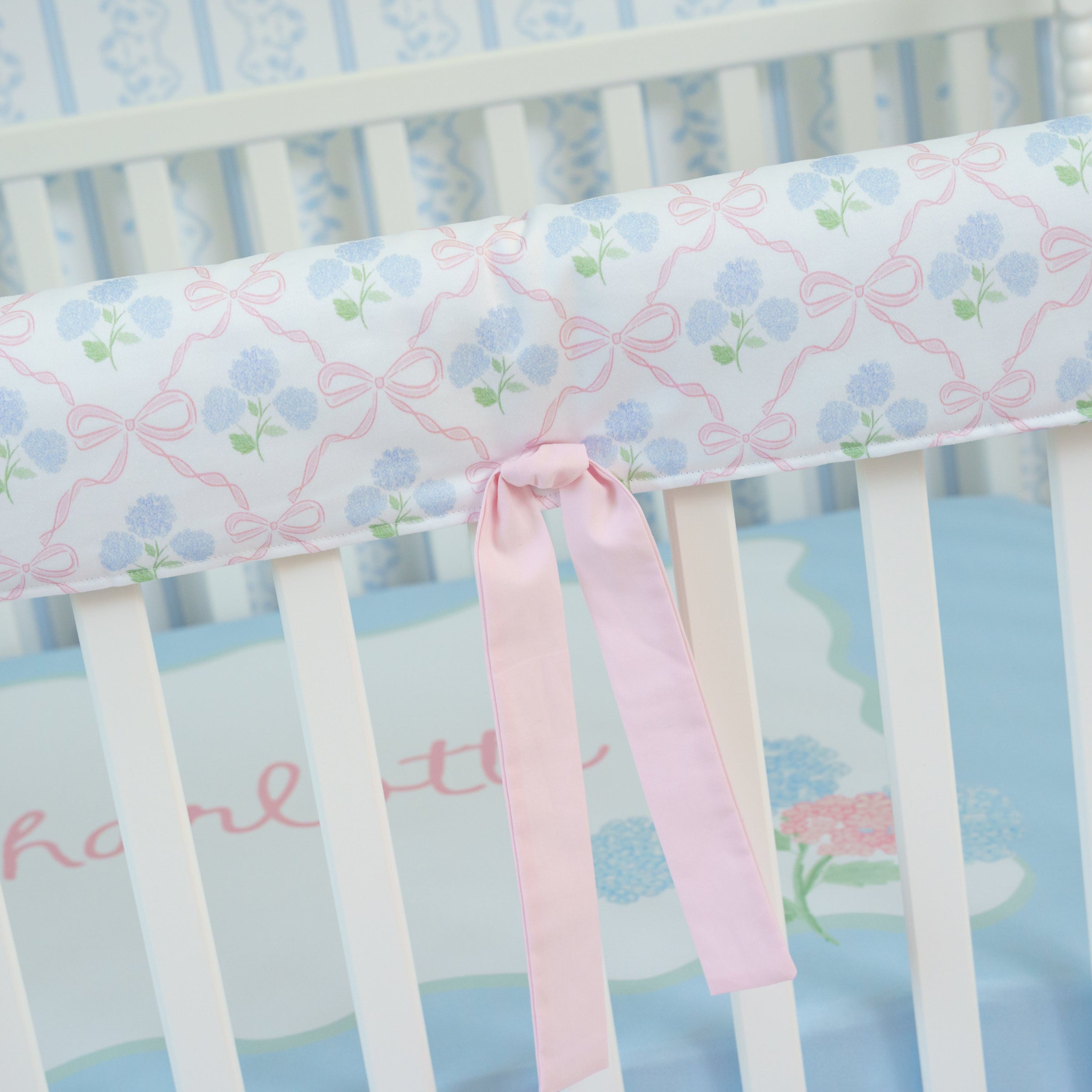 Crib with floral-patterned bumper and pink ribbon, on a light blue background