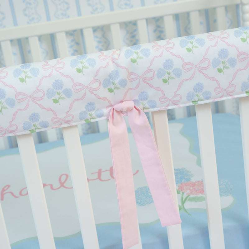 Crib with floral-patterned bumper and pink ribbon, on a light blue background