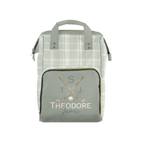 A personalized golf-themed diaper bag featuring a light gray and white plaid body, a solid sage green front pocket with gold zipper trim, and a custom monogram print reading 'S T J THEODORE James' with crossed golf clubs and a golf ball.