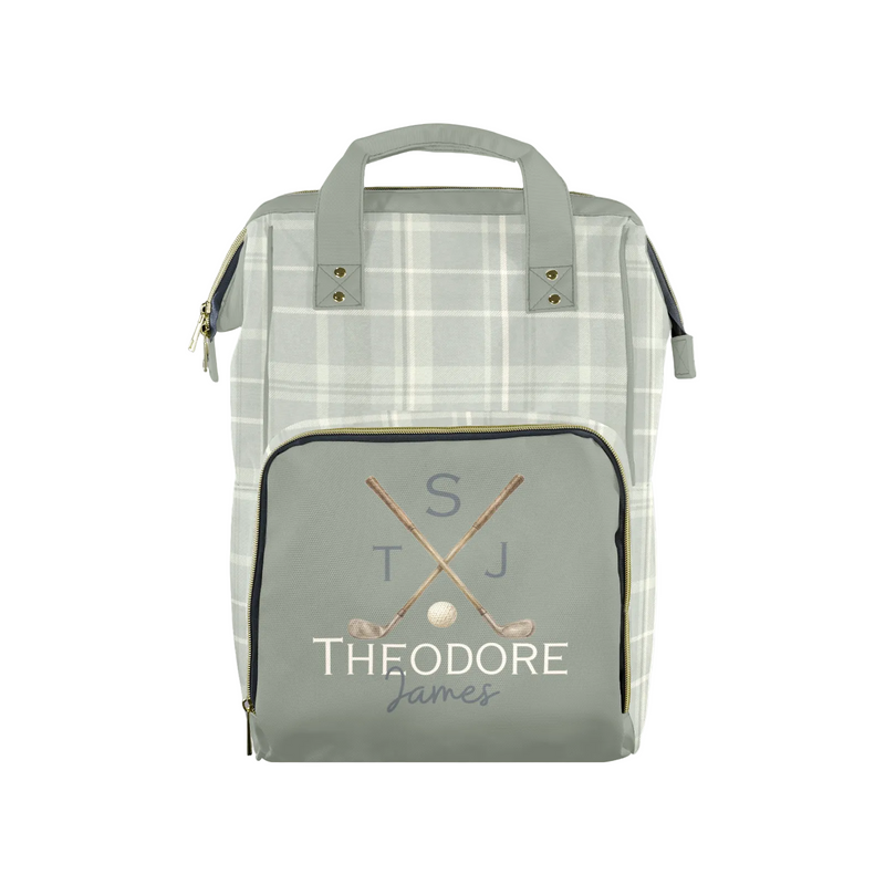 A personalized golf-themed diaper bag featuring a light gray and white plaid body, a solid sage green front pocket with gold zipper trim, and a custom monogram print reading 'S T J THEODORE James' with crossed golf clubs and a golf ball.