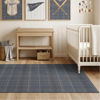 A styled nursery room featuring a large slate blue plaid area rug on a light wood floor, with a white crib, changing table, and armchair. Three framed baseball-themed wall art prints hang above the changing table.
