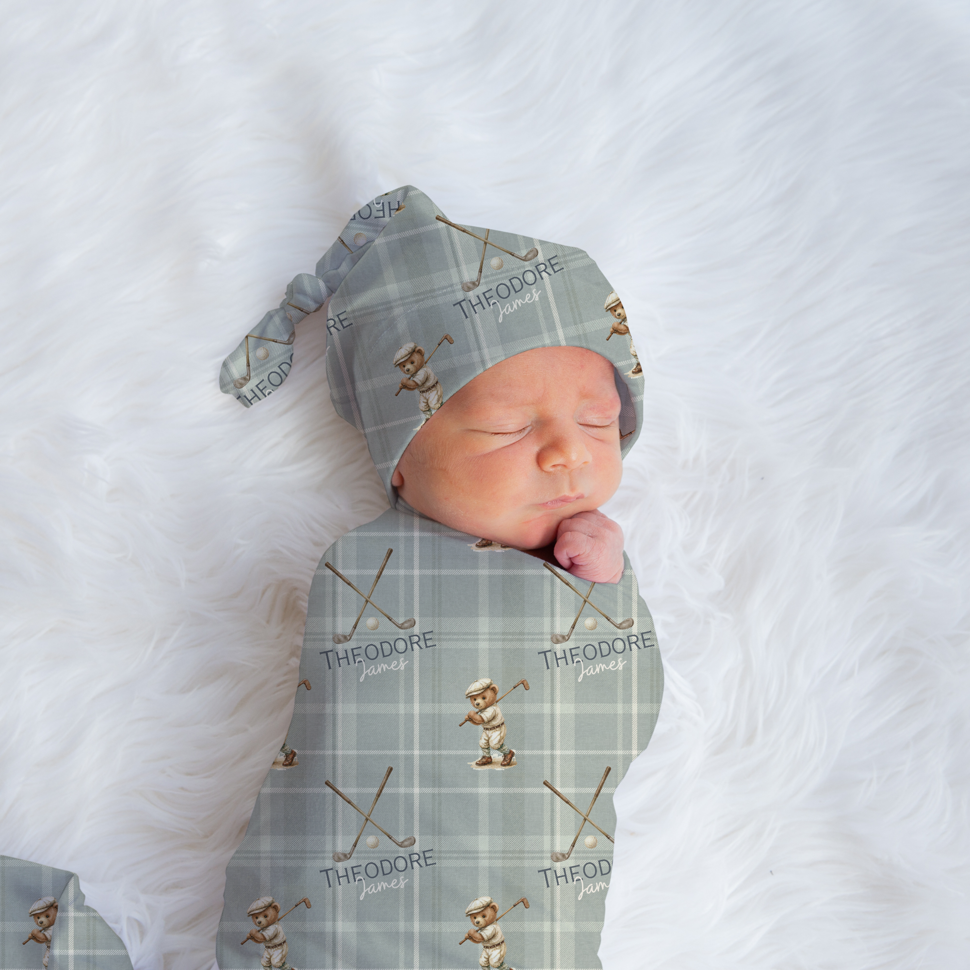 A sleeping newborn baby swaddled and bonneted in a sage green plaid fabric with a small teddy bear and crossed skates print, lying on a white faux fur background.