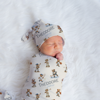 A sleeping newborn baby swaddled and wearing a matching printed swaddle wrap and hat set, laid on a white faux fur blanket. The set features a cream background with a repeating pattern of small cartoon bears and the name 'THEODORE' printed in black.