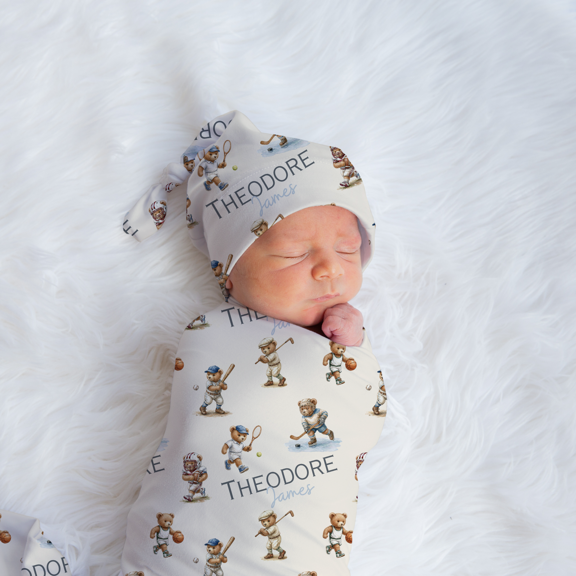 A sleeping newborn baby swaddled and wearing a matching printed swaddle wrap and hat set, laid on a white faux fur blanket. The set features a cream background with a repeating pattern of small cartoon bears and the name 'THEODORE' printed in black.